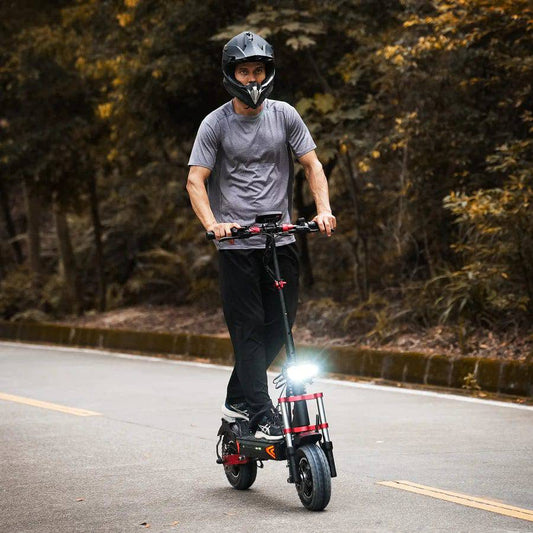 A man riding a high-performance electric scooter on a paved road surrounded by autumn foliage, wearing a black protective helmet and casual attire. The scooter is equipped with bright front LED lights for visibility.